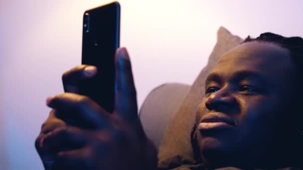 Молодой африканский американец проверяет свой смартфон с улыбкой, расслабляясь на диване — стоковое видео