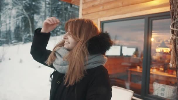 Mulher bonita ajustando seu cabelo longo na frente da casa de montanha de madeira e apreciando a queda de neve — Vídeo de Stock
