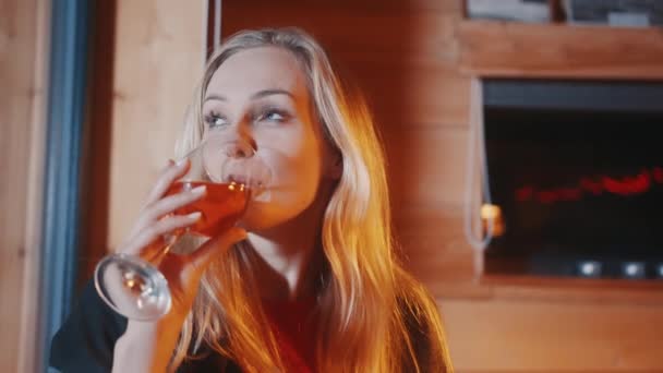 Ευτυχισμένη νεαρή γυναίκα που πίνει από ένα ποτήρι κρασί και κοιτάζει μέσα από το παράθυρο το χειμώνα — Αρχείο Βίντεο