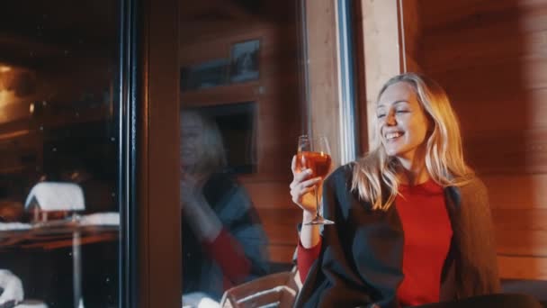 Jonge vrouw die warme thee drinkt met citroen uit het wijnglas. Zittend bij het raam en kijkend naar sneeuw — Stockvideo
