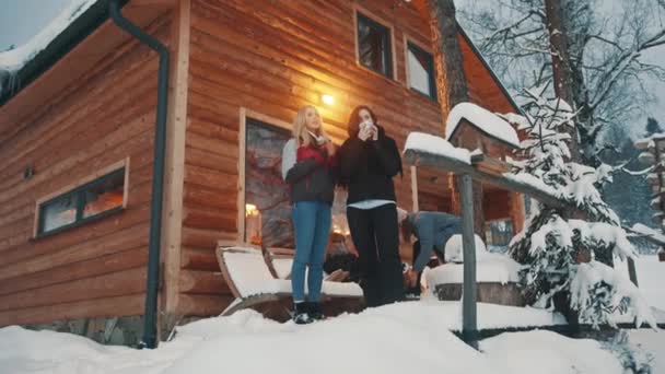 Amigos passando férias de inverno na montanha. Beber bebida quente na frente da casa de madeira e desfrutar de neve — Vídeo de Stock