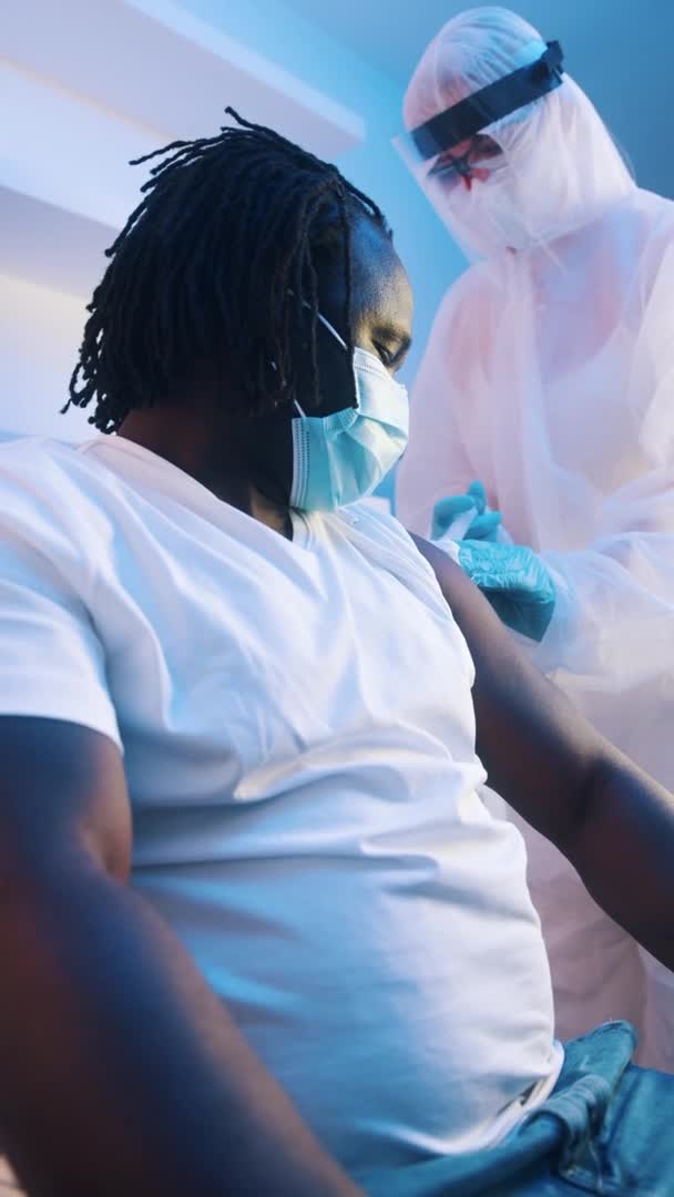 По вертикали. Медсестра в защитном костюме дает вакцину против коронавируса африканскому чернокожему мужчине — стоковое видео