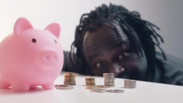 Decisión financiera, inversiones y ahorros. Hombre americano africano divertido mirando en las monedas en frente de la alcancía. — Vídeos de Stock