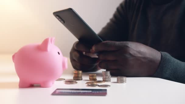 Mannen som använder telefon för att kolla sitt bankkonto. Piggy bank kreditkort och mynt på bordet. Investeringar med hjälp av mobilappen — Stockvideo