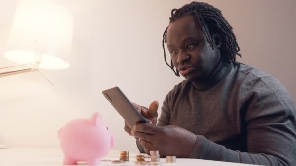 Mannen som använder telefon för att kolla sitt bankkonto. Piggy bank kreditkort och mynt på bordet. Investeringar med hjälp av mobilappen — Stockvideo