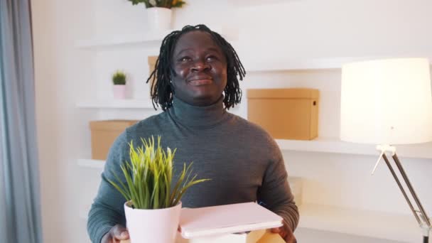 Feliz joven negro reubicándose en el nuevo apartamento. Cajas de cartón con plantas y libros. — Vídeos de Stock