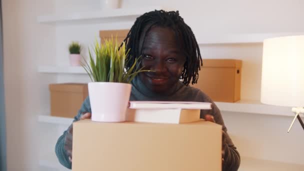 興奮した若いアフリカ系アメリカ人の男性の肖像画は、新しいアパートに移動します。植物や書籍と段ボール箱を保持 — ストック動画