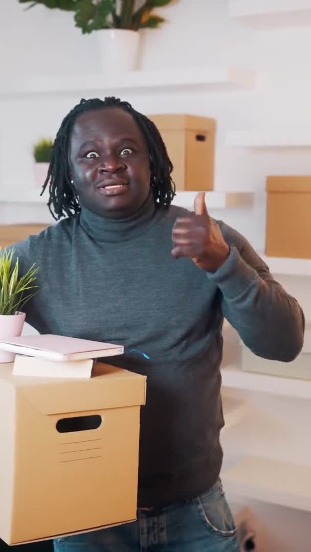 Verical. Black man moving in to the new apartment. Holding cardboard boxes with plant and books. Thumbs up — Stock Video