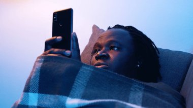 Kanepede battaniyeyle kaplı bir şekilde sosyal medyayı kontrol eden genç bir zenci.