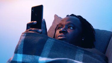 Afro-amerikan yorgun siyah adam kanepede yatarken akıllı telefon tutuyor. 