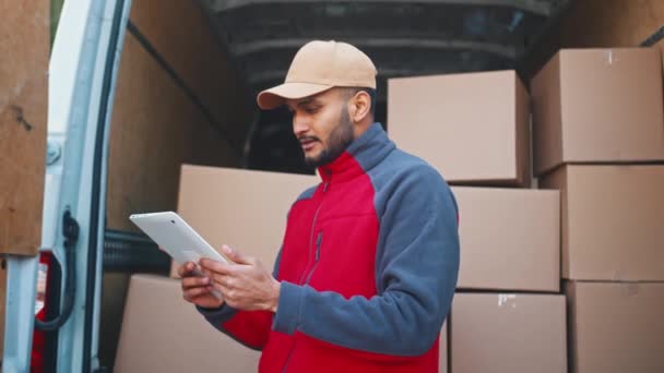 Entrega hombre utilizando la tableta para obtener una vista previa de pedidos. De pie frente a la furgoneta con paquetes — Vídeos de Stock