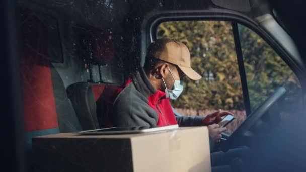 Kurier kontaktuje się z klientem za pomocą smartfona na siedzeniu kierowcy. Mężczyzna z maską dostarczającą opakowanie — Wideo stockowe