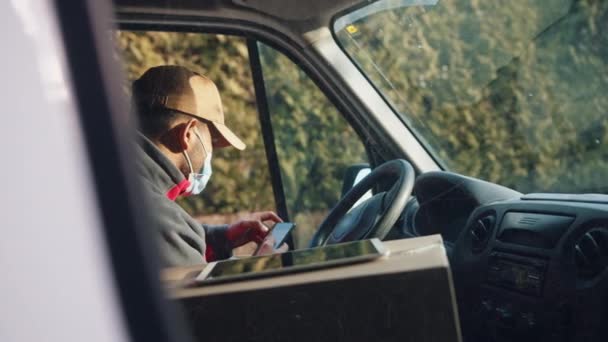 坐在驾驶座上使用智能手机的信使。配戴口罩的送货人 — 图库视频影像