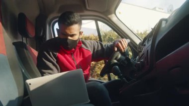 Genç adam dizüstü bilgisayara bakıyor ve minibüsün motorunu çalıştırıyor.