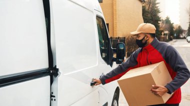 Yüzü maskeli genç minibüsten bir paket alıyor ve kapıyı kapatıyor. Covid19 sırasında teslimat