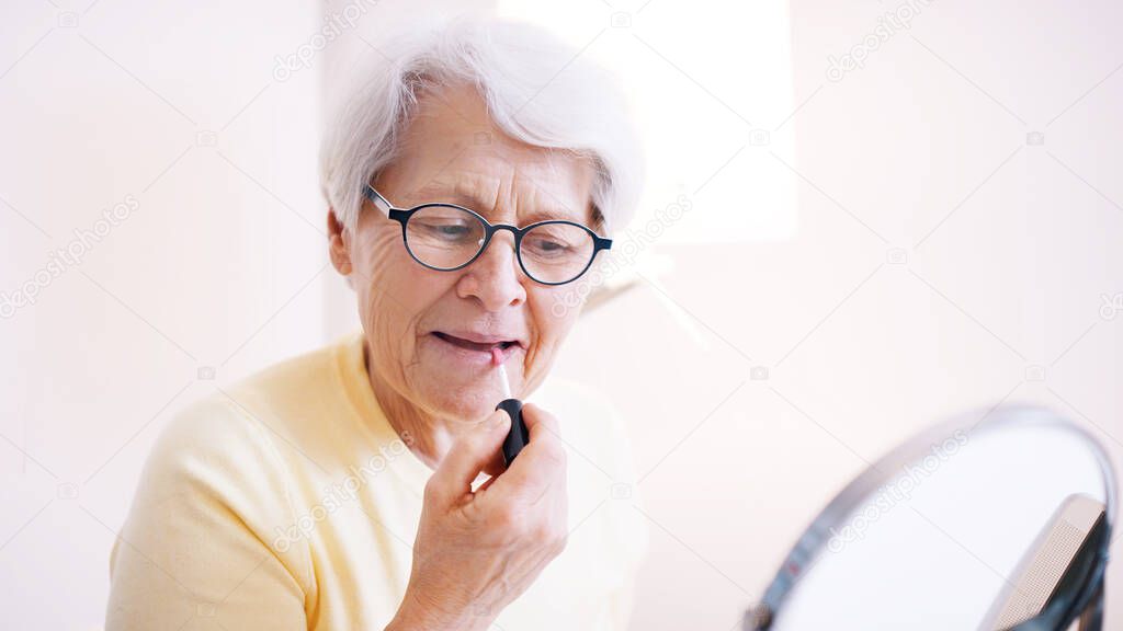 Elderly woman applying lip glow. Refflection in the mirror