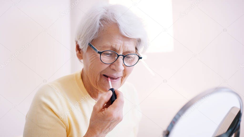 Elderly woman applying lip glow. Refflection in the mirror
