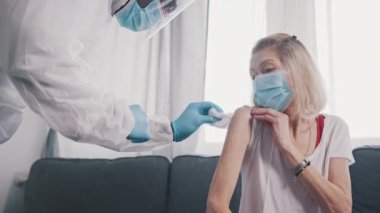 Koruyucu kıyafetli hemşire yaşlı bir kadının koluna kovid aşısı enjekte ediyor.