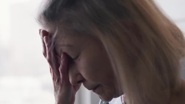 Vieja mujer solitaria que sufre de dolor de cabeza. Mirando por la ventana — Vídeo de stock