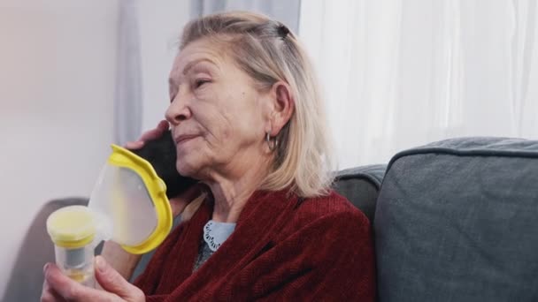 Пожилая женщина использует ингалятор кислорода во время телефонного разговора. Уязвимый человек во время вспышки ковидов — стоковое видео