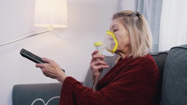 Mujer anciana usando máscara de oxígeno mientras cambia canales en la televisión en casa — Vídeo de stock