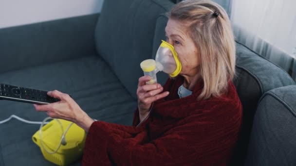 Mujer anciana usando máscara de oxígeno mientras cambia canales en la televisión en casa — Vídeo de stock