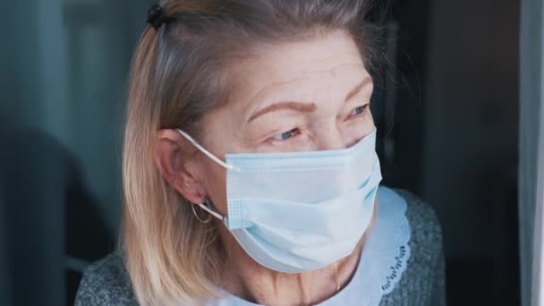 Portrait of elderly woman with medical face mask in quarantine looking through the window — Stock Video