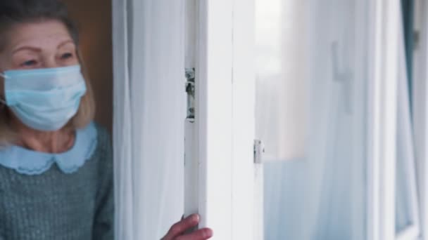 Oudere grijze vrouw met medisch masker die door het raam kijkt — Stockvideo