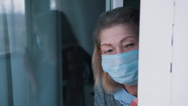 Πορτρέτο της μοναχικής ηλικιωμένης γυναίκας σε καραντίνα με μάσκα προσώπου κοιτάζοντας μέσα από το παράθυρο — Αρχείο Βίντεο