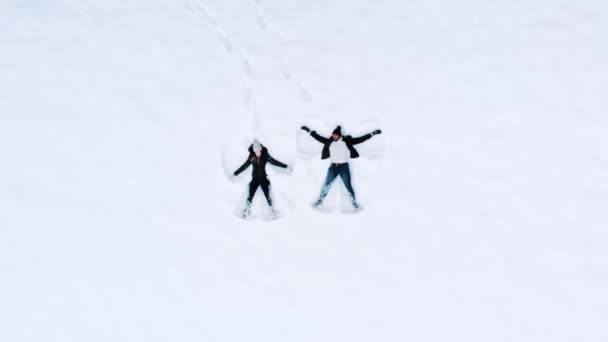 Jovem casal feliz deitado de volta na neve durante o inverno em movimento braços e pernas — Vídeo de Stock