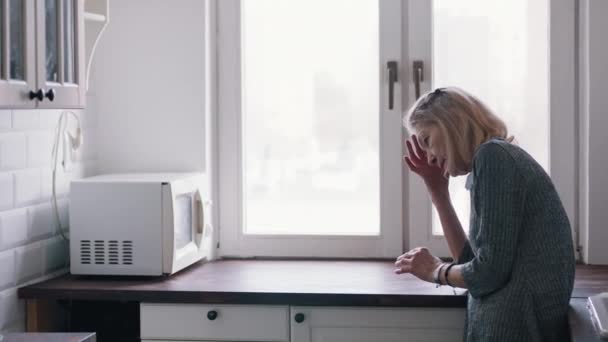 Dolorosa anciana solitaria apoyada en el mostrador de la cocina y mirando por la ventana — Vídeos de Stock