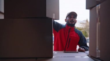 Genç Hintli adam teslimat siparişini kontrol etmek için tablet kullanıyor. Karton kutularla kamyon yükleniyor
