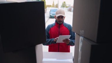 Genç Hintli adam teslimat siparişini kontrol etmek için tablet kullanıyor. Karton kutularla kamyon yükleniyor