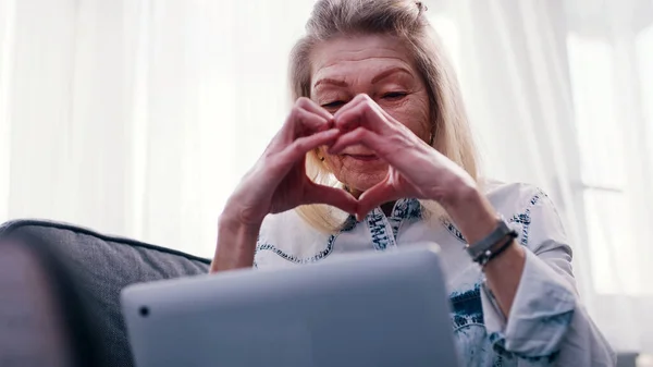 Una anciana que usa un portátil para una videollamada. Enviando corazón a su familia — Foto de Stock