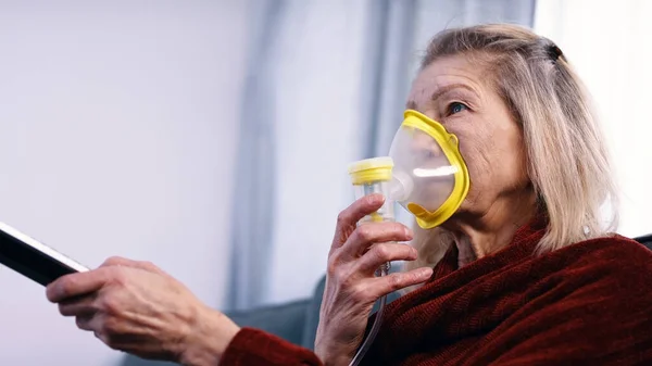 Уязвимая старуха в карантине, использующая ингалятор кислорода и меняющая каналы на телевидении — стоковое фото