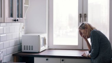 Mutfak tezgahına yaslanıp pencereden bakan zayıf, yalnız, yaşlı kadın.