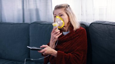 Evde televizyon kanallarını değiştirirken oksijen maskesi kullanan yaşlı bir kadın.