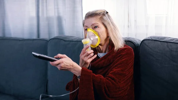 Пожилая женщина использует кислородную маску при переключении каналов на ТВ дома — стоковое фото