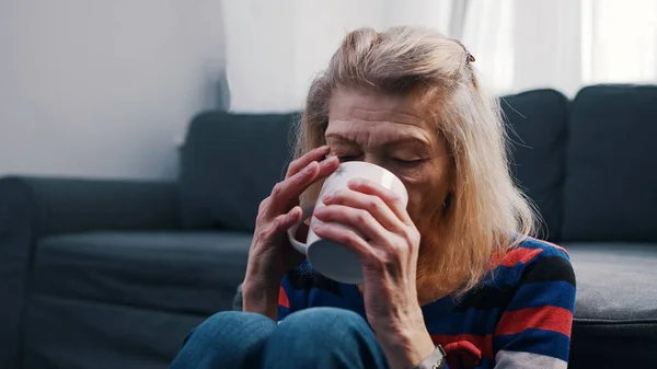 Одинокая старуха пьет горячий напиток, сидя на полу перед диваном — стоковое фото