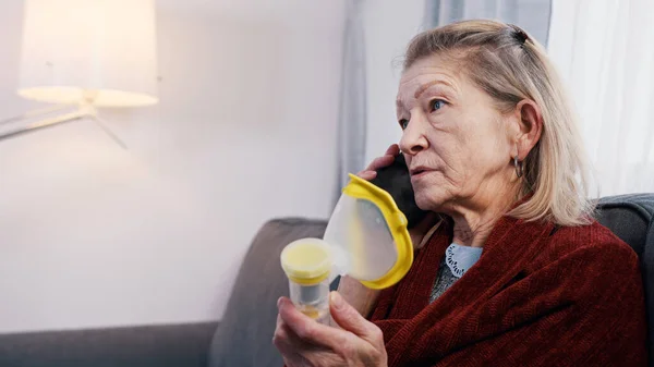Пожилая женщина использует ингалятор кислорода во время телефонного разговора. Уязвимый человек во время вспышки ковидов — стоковое фото