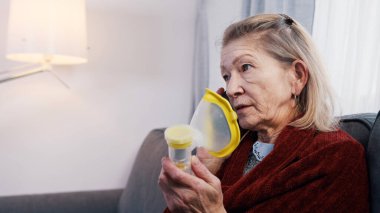 Savunmasız yaşlı kadın telefonla konuşuyor ve astım spreyi kullanıyor. Yaşlandıkça nefes almakta güçlük çekiyorum.