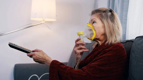 Mujer anciana usando máscara de oxígeno mientras cambia canales en la televisión en casa — Foto de Stock