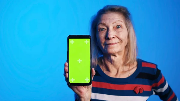 Mujer de edad con los ojos bien abiertos sosteniendo teléfono inteligente con pantalla verde — Foto de Stock