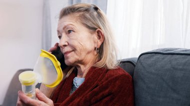 Savunmasız yaşlı kadın telefonla konuşuyor ve astım spreyi kullanıyor. Yaşlandıkça nefes almakta güçlük çekiyorum.