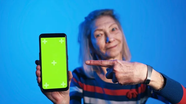 Starší žena drží smartphone se svislou zelenou obrazovkou a s úsměvem na něj ukazuje prstem — Stock fotografie