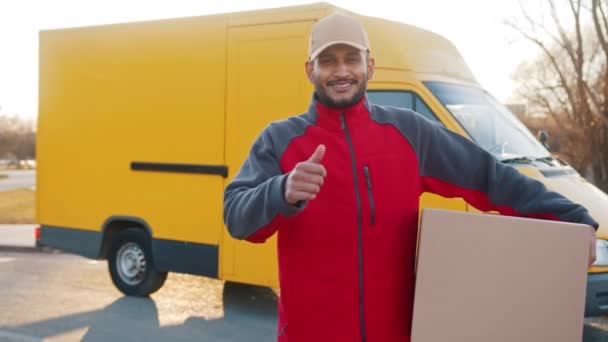 Smiling adult delivery man carrying cardboard box, showing thumb up gesture and looking at camera. Parcel delivery from eshop concept. — Stock Video