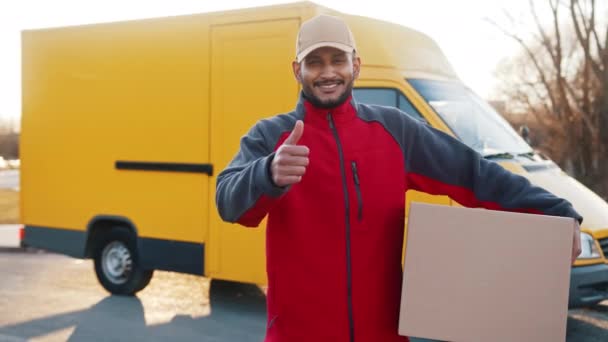 Smiling adult delivery man carrying cardboard box, showing thumb up gesture and looking at camera. Parcel delivery from eshop concept. — Stock Video