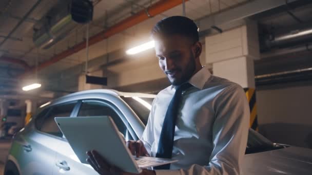 Vue de la taille de souriant homme d'affaires adulte debout à la voiture dans le parking souterrain. Regarder un ordinateur portable et faire un geste de succès. — Video