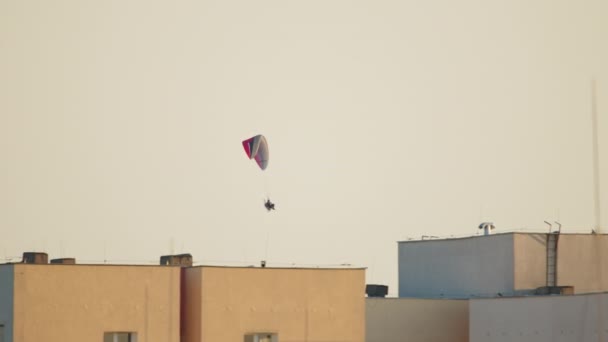 Paraşütçü, paraşütün altındaki binaların üzerinde uçuyor. Temiz beyaz bir gökyüzü. — Stok video