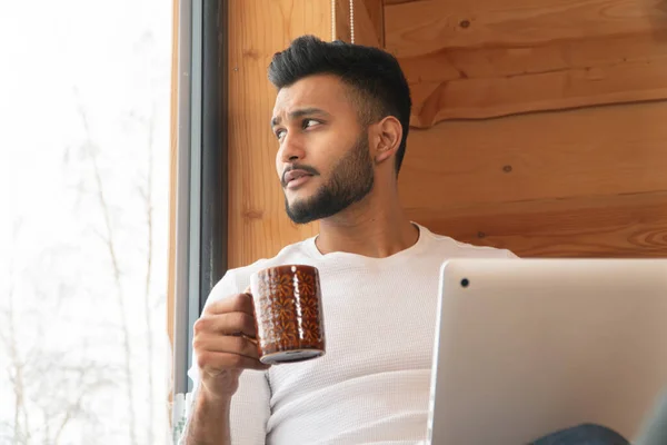 Ένας Ινδιάνος κρατάει μια κούπα του καφέ δίπλα στο παράθυρο με το λάπτοπ του. — Φωτογραφία Αρχείου